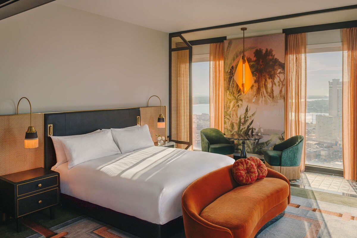 Habitación de hotel con una cama grande, una mesita de noche, un sillón verde, una pequeña mesa con una lámpara y vistas a la ciudad a través de grandes ventanales.