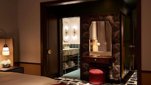 A hotel room with a bed, a nightstand, a mirror, a vanity with a sink, and a red ottoman.