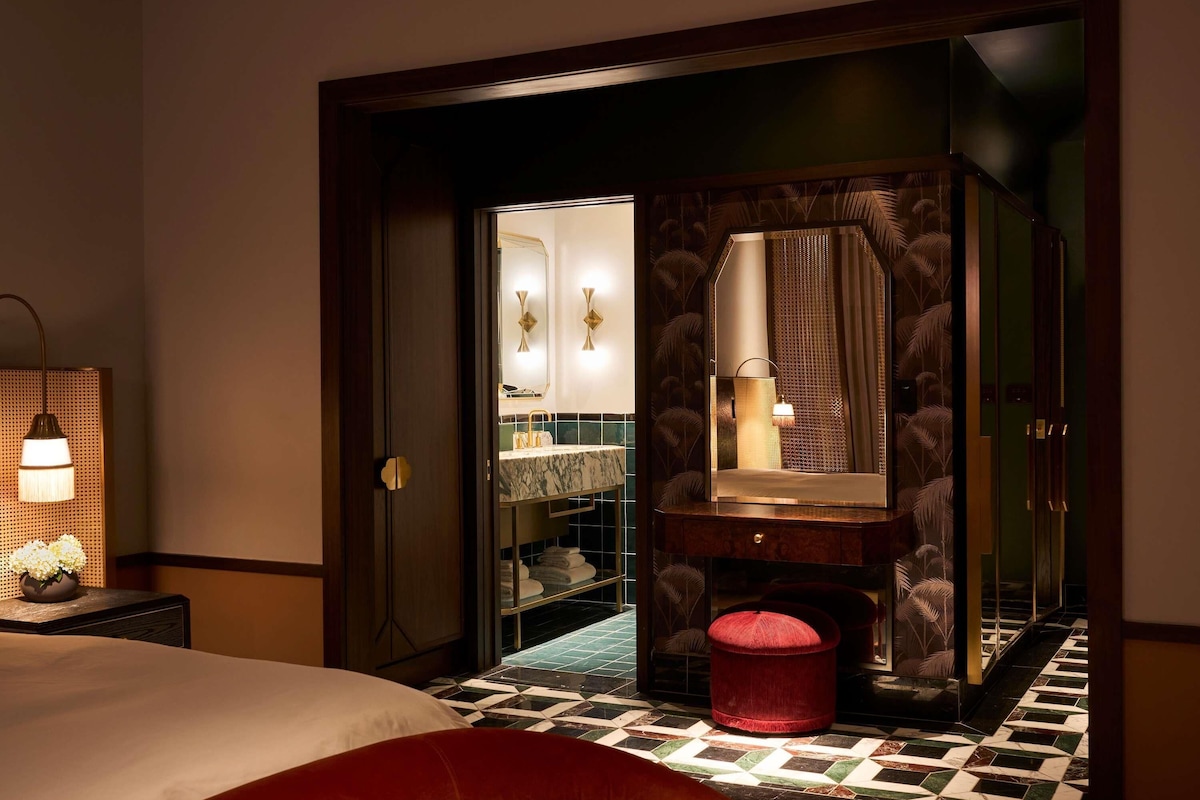 A hotel room with a bed, a nightstand, a mirror, a vanity with a sink, and a red ottoman.