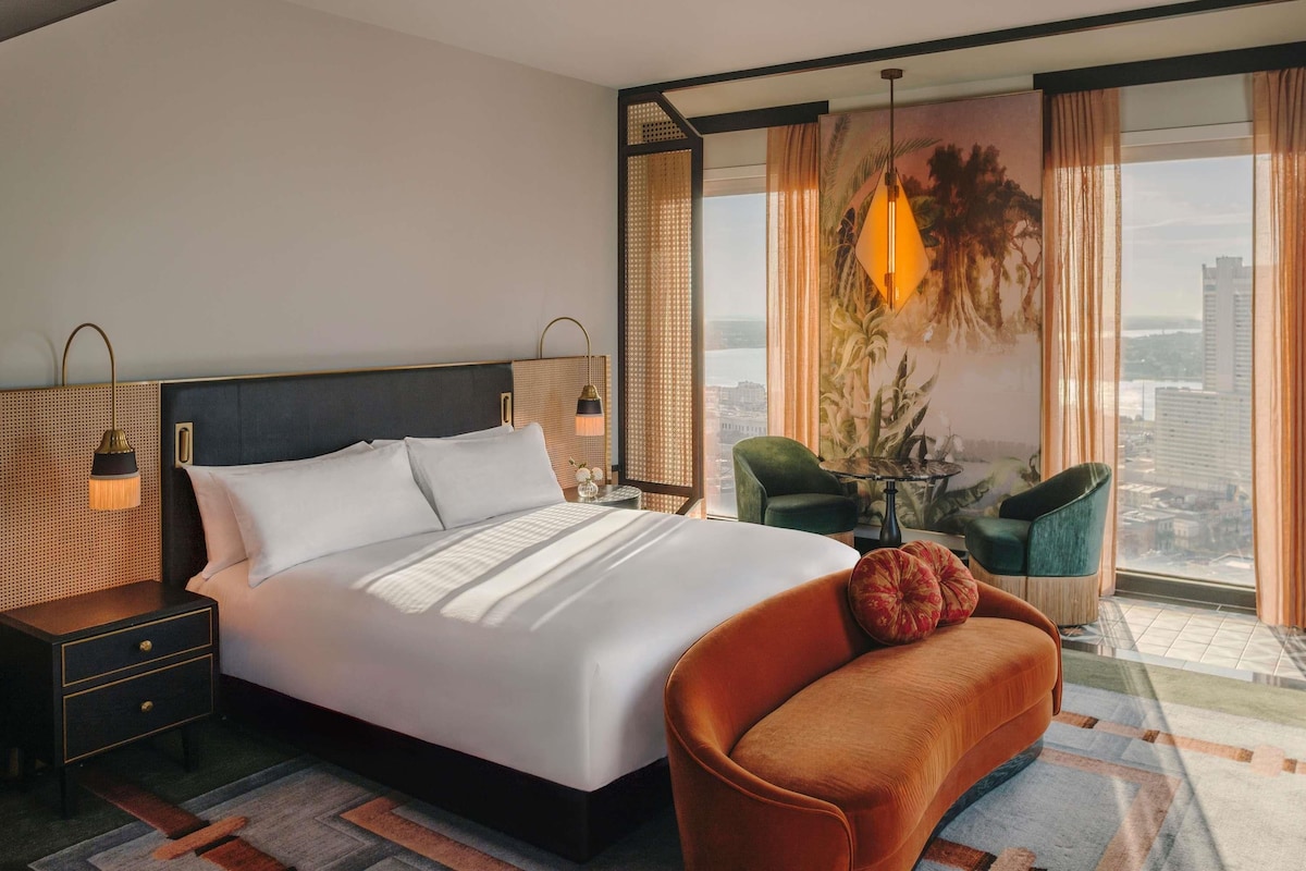 A hotel room with a large bed, a nightstand, a green armchair, a table with a lamp, and a view of the city through large windows.