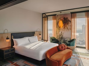 A hotel room with a large bed, a nightstand, a green armchair, a table with a lamp, and a view of the city through large windows. - Fairmont New Orleans (Opening November 2026) (New Orleans)