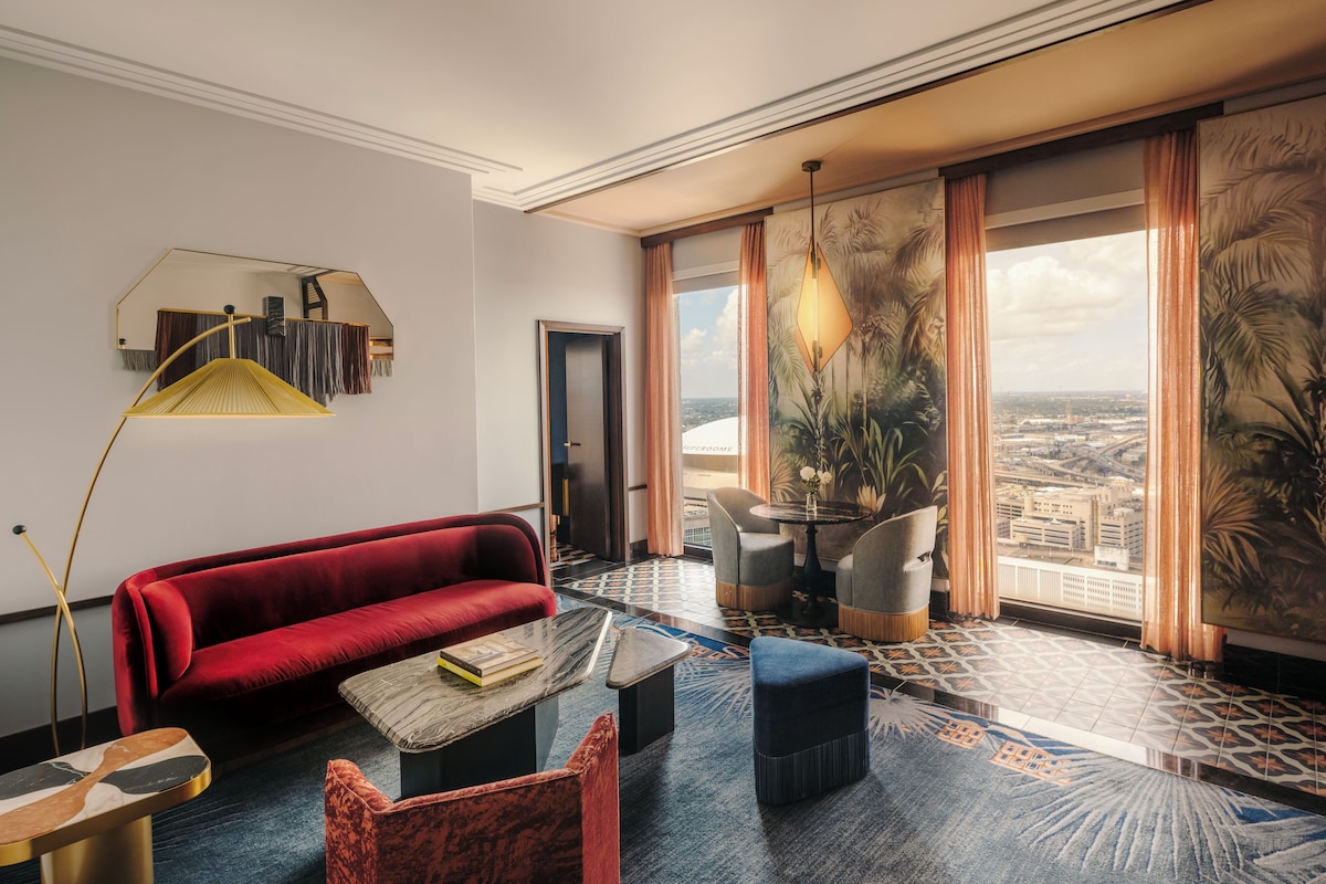 A modern hotel room with a red sofa, a coffee table, a dining area with chairs, and a large window offering a city view.