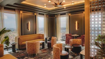 A modern hotel room with a large window, a sofa, a dining table, and multiple chairs.