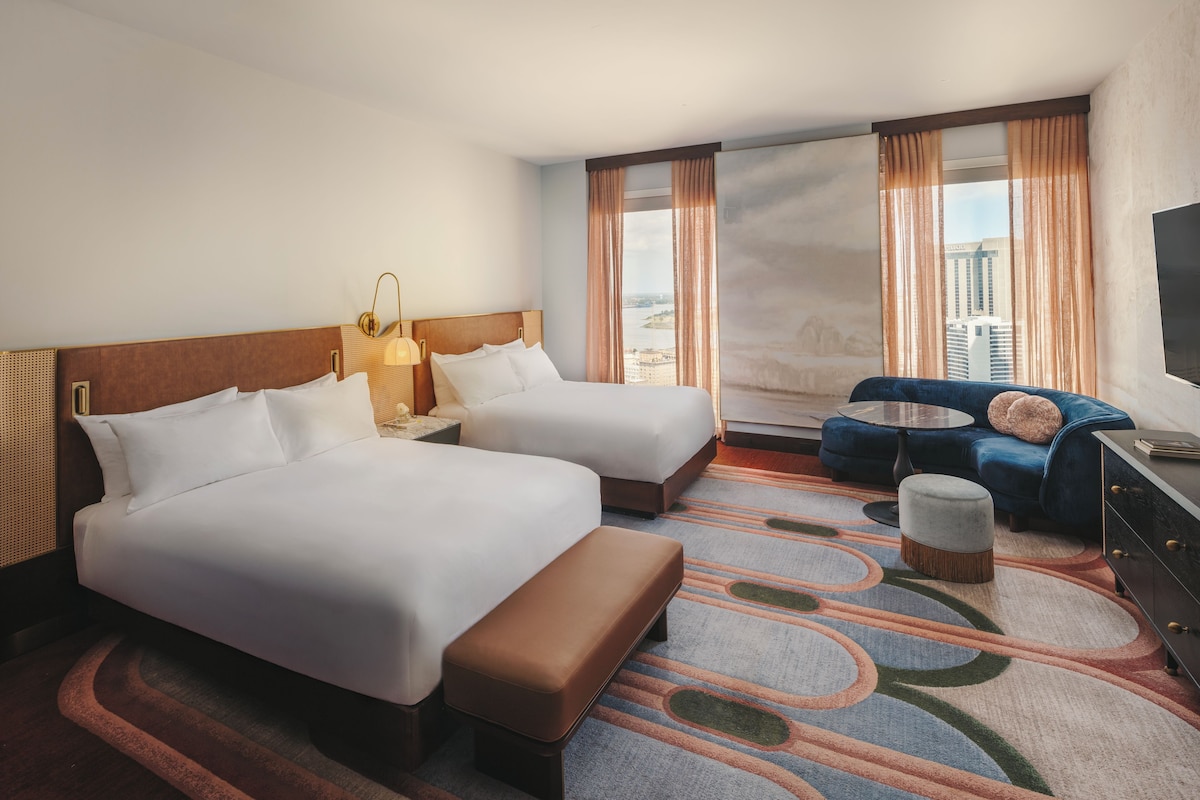 A hotel room with two beds, a sofa, a TV, and a view of a cityscape.