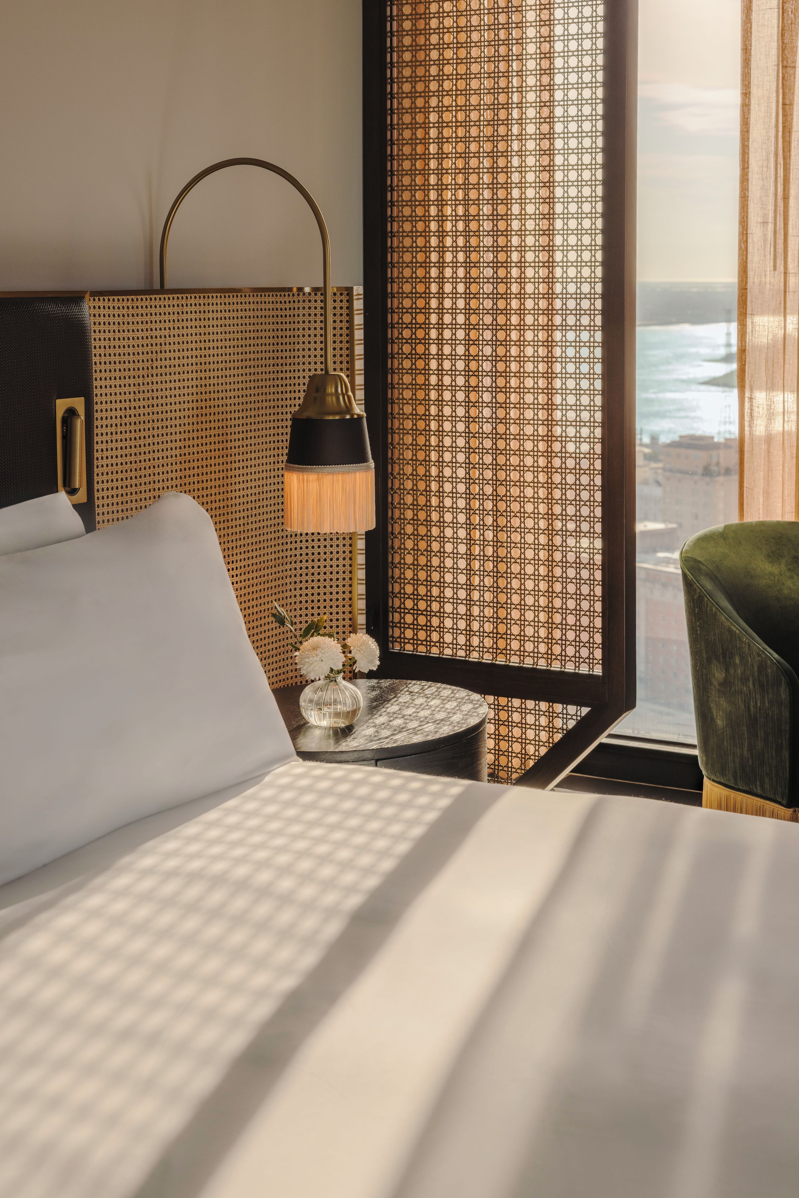 Quarto de hotel com cama, mesa de cabeceira, abajur, vaso com flores e vista para o mar pela janela.