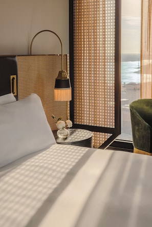 Una habitación de hotel con cama, mesita de noche, lámpara, un jarrón con flores y vista al océano a través de una ventana.