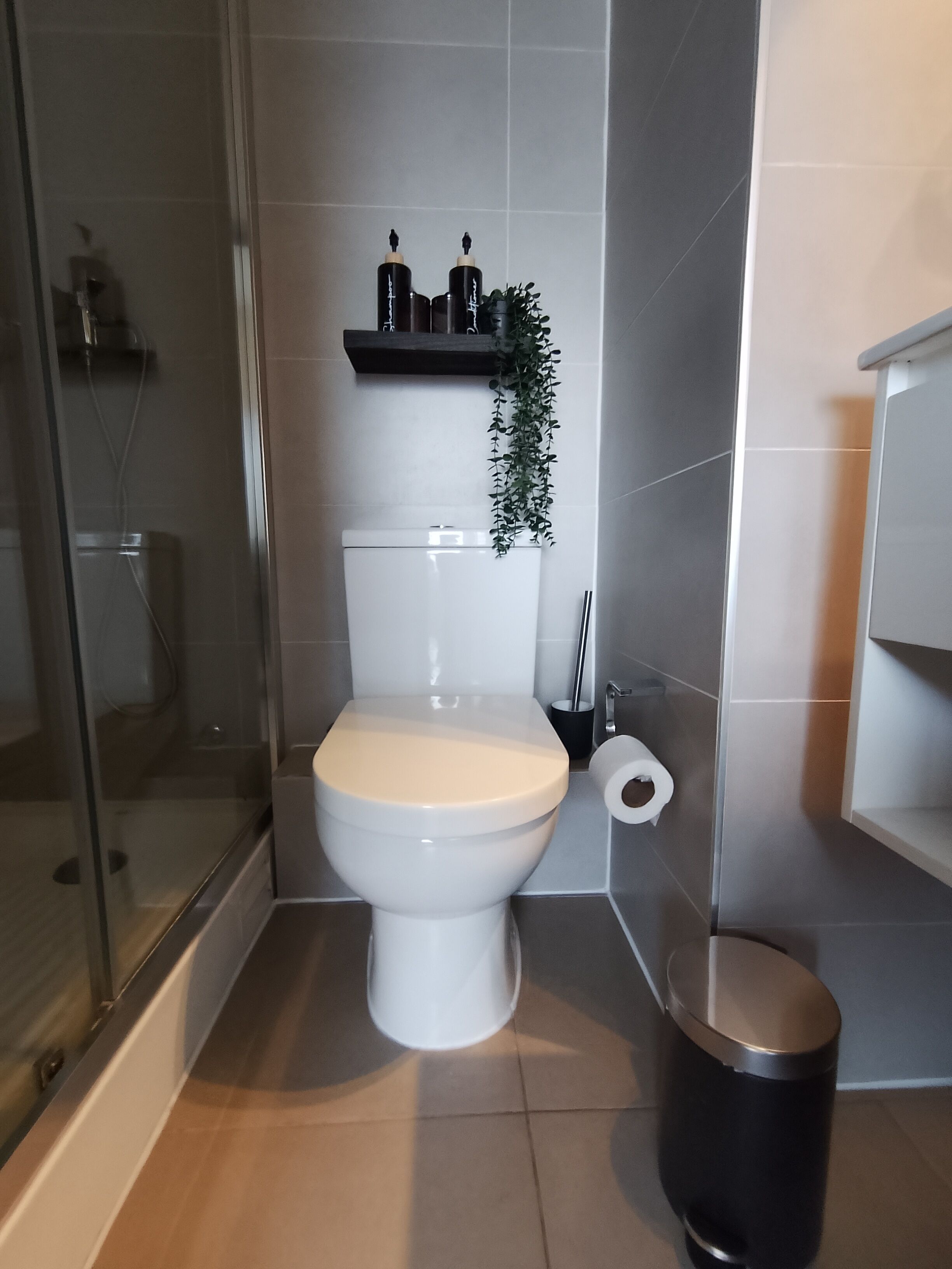 Et badeværelse med toilet, glasvæg omkring bruseren, skraldespand og et hyldesæt med toiletartikler.