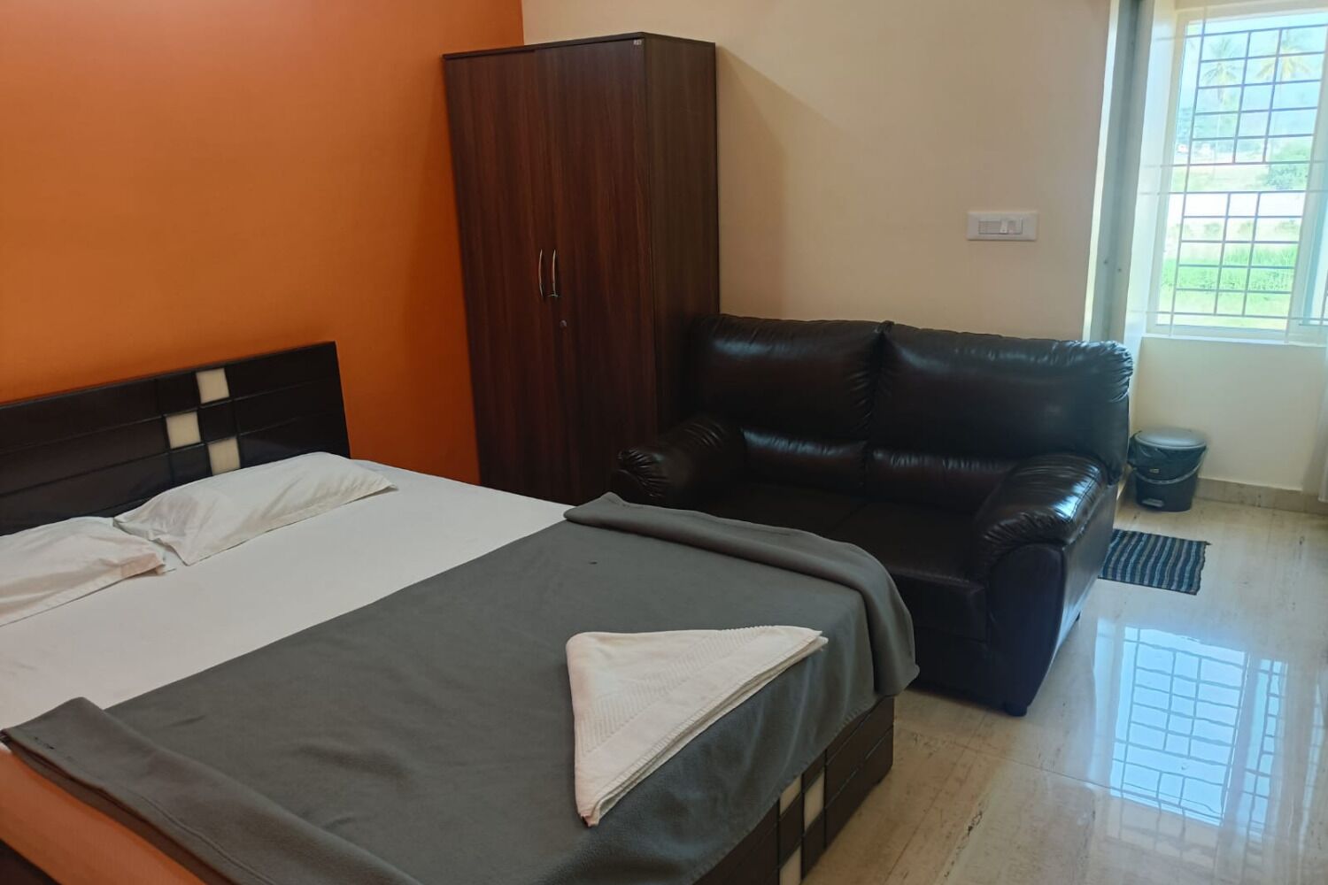 Un dormitorio con cama, armario, sofá de cuero, ventana y un cubo de basura.