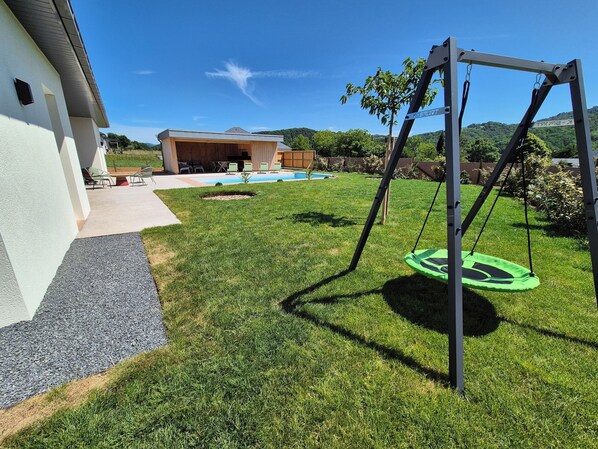 A backyard with a swing set, a pool, and a covered patio area.