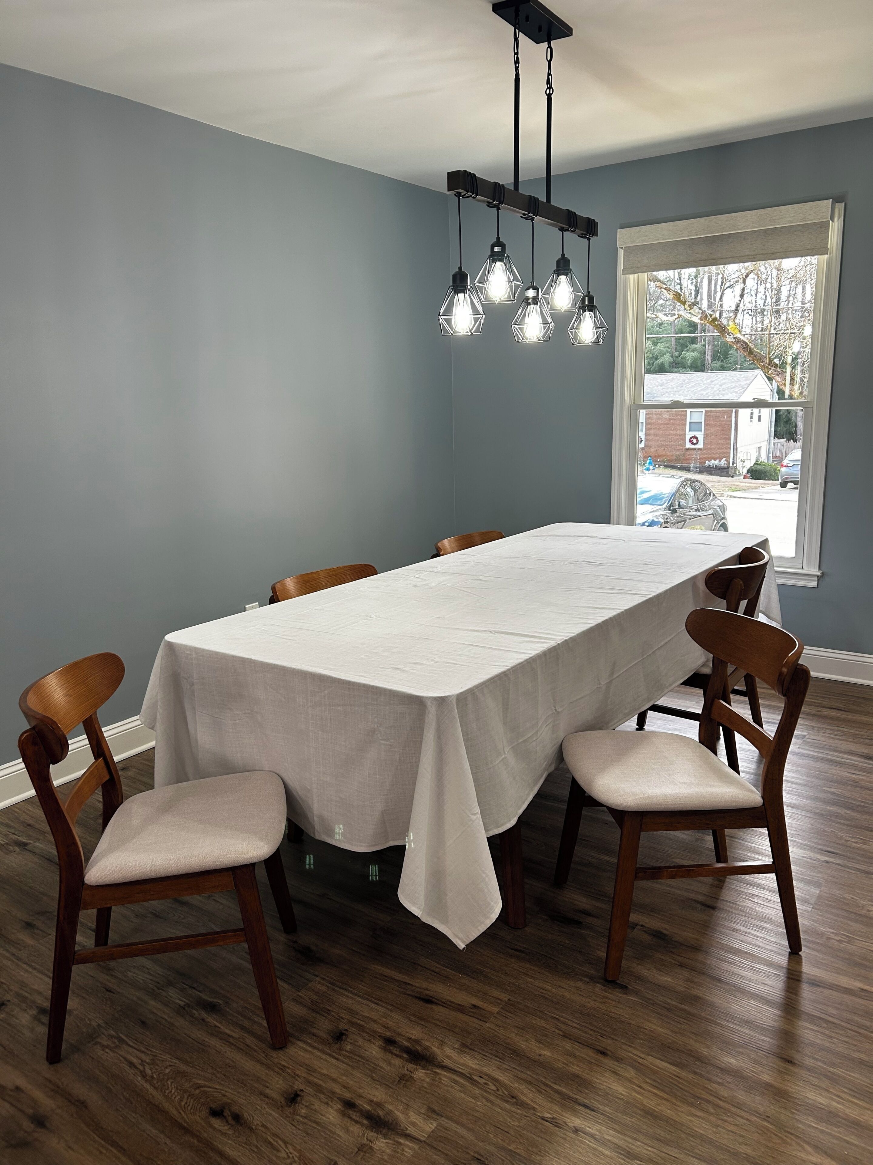 Ein Esszimmer mit einem langen Tisch, sechs Holzsesseln, einer weißen Tischdecke und einer hängenden Leuchte.