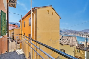 Un edificio amarillo con un balcón, barandilla metálica y vistas al agua y montañas a lo lejos.