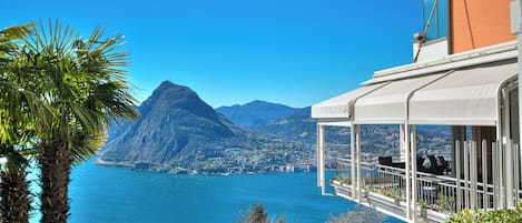 A coastal view with a mountain, a building with a balcony overlooking the sea, and lush greenery.