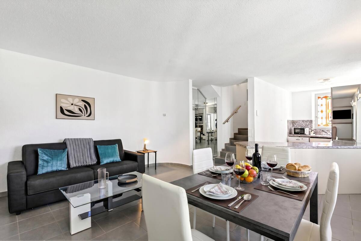 Una sala de estar moderna con comedor, un sofá de cuero negro, una mesa de centro de vidrio y una cocina visible a través de una puerta abierta.