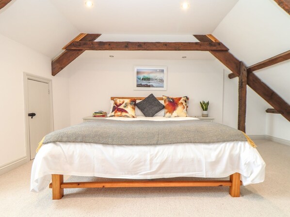 Un dormitorio con cabecera de cama de madera, ropa de cama blanca, una mesita de noche con libros, una planta en maceta y un cuadro en la pared.