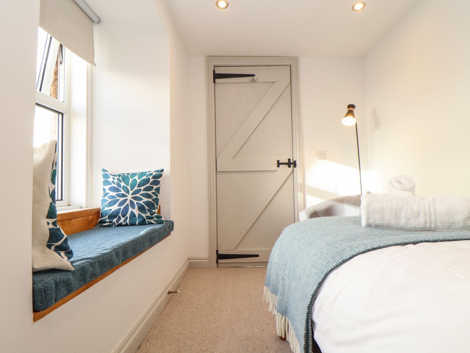 Un dormitorio con una cama, un rincón con asiento junto a la ventana con un cojín, una puerta y una lámpara de pie.