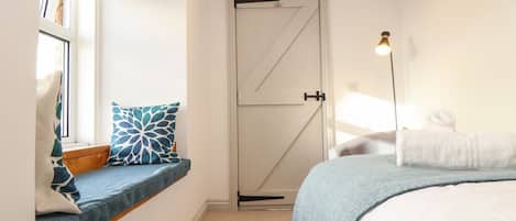 Un dormitorio con una cama, un rincón con asiento junto a la ventana con un cojín, una puerta y una lámpara de pie.