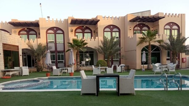 A pool area with lounge chairs, a table, and a building with arched windows and a castle-like facade.