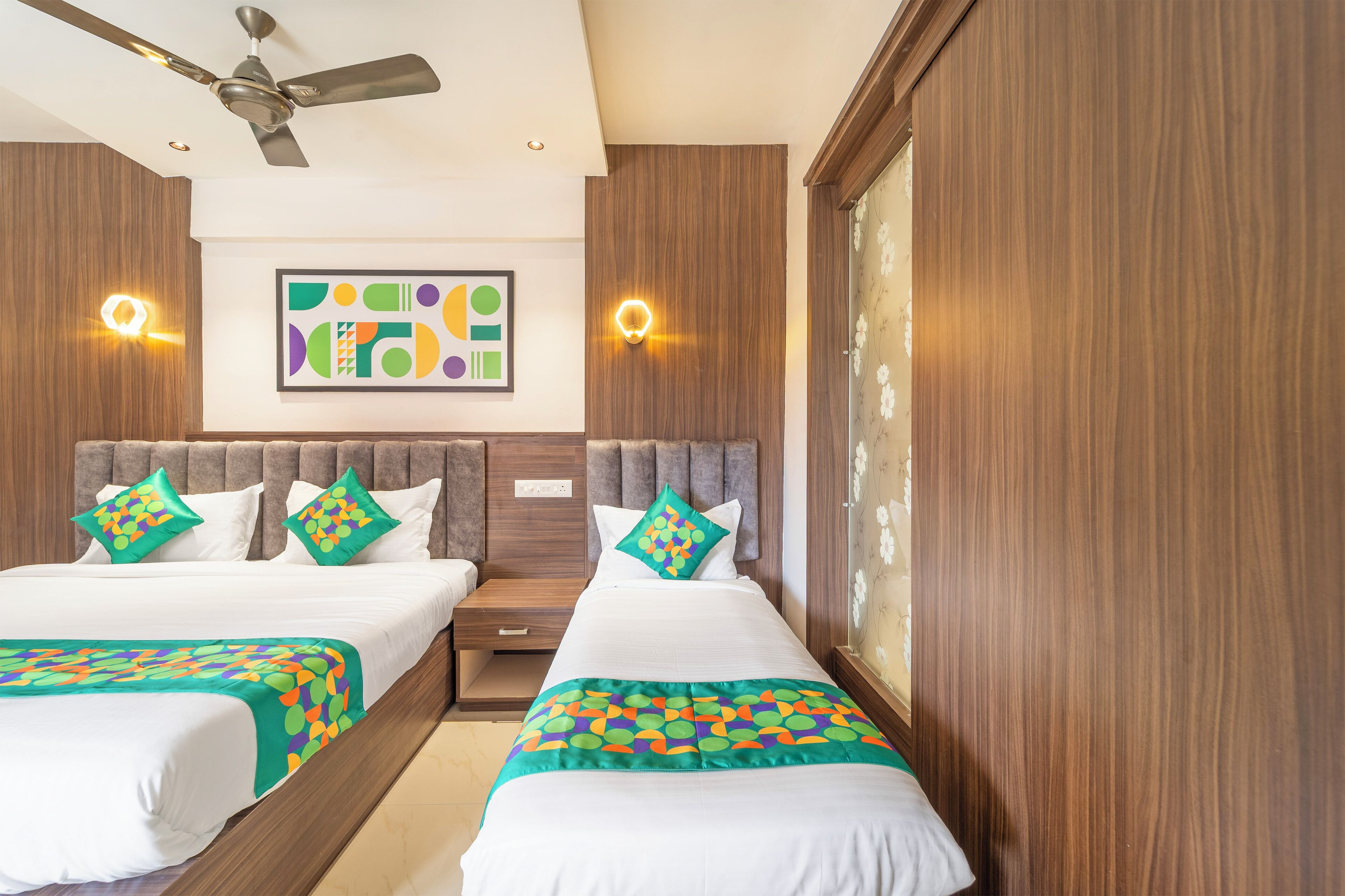 A hotel room with two beds, a ceiling fan, a wall-mounted artwork, and a window with a floral pattern.
