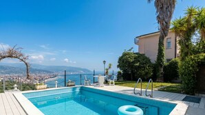 Ein rechteckiger Pool mit glasklar blauem Wasser, umgeben von einer gefliesten Liegefläche, einem Liegestuhl, einem kleinen Tisch und Blick auf eine Küstenstadt.