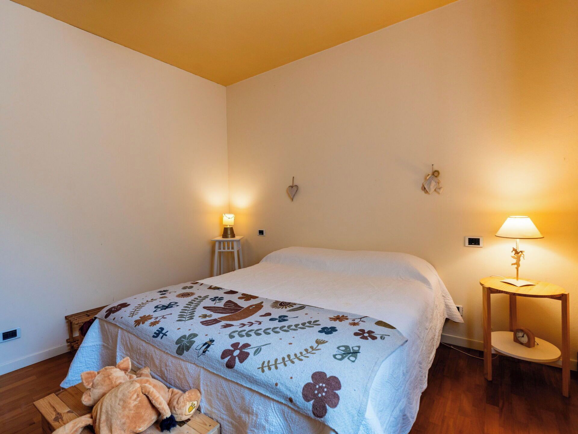 A bedroom with a bed, two bedside lamps, a small table with a lamp, a teddy bear, and a wall clock.
