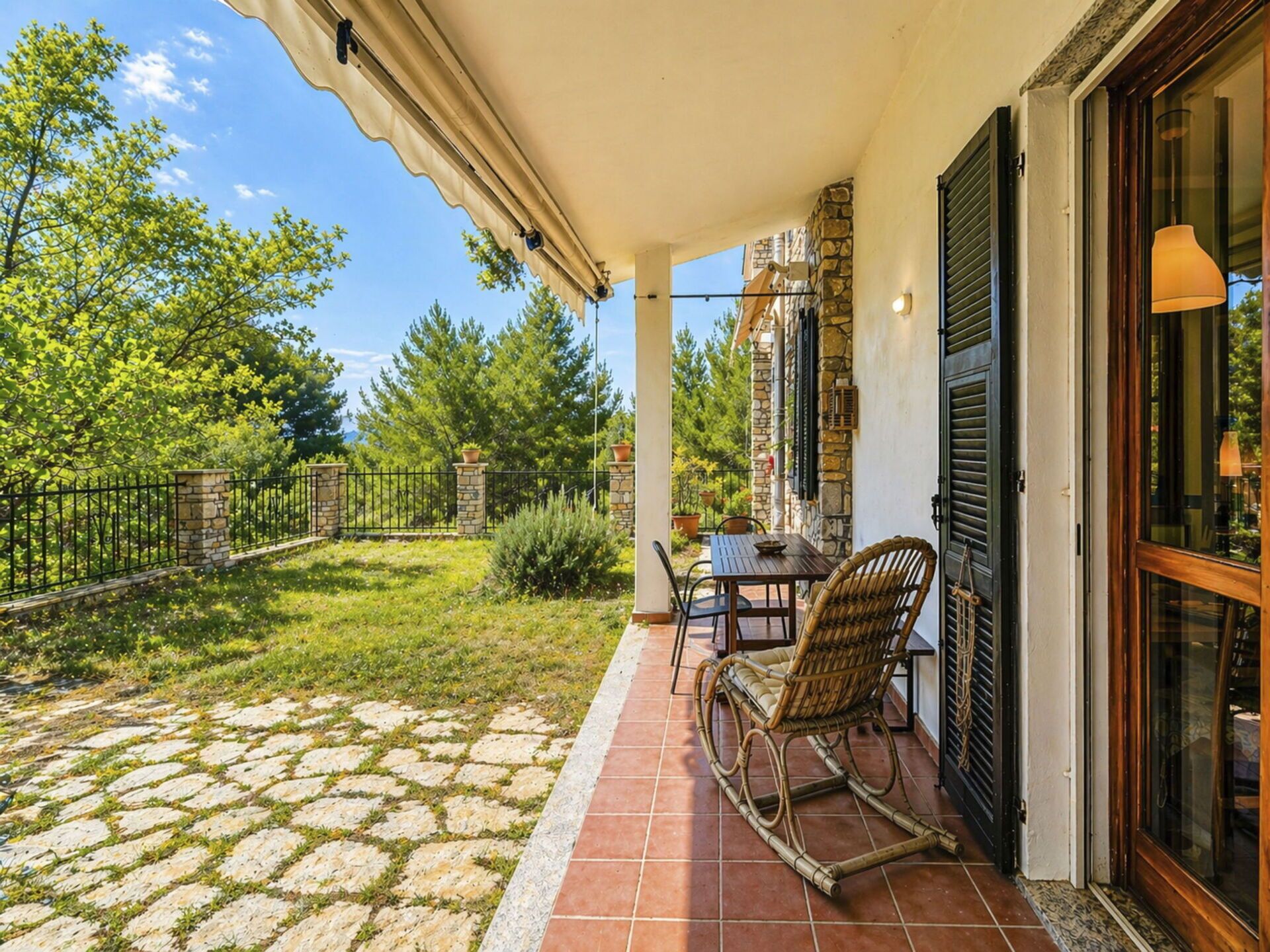 A covered patio with a table and chairs, a stone wall, and a view of greenery.