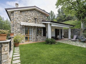 Ein Steinhäuschen mit überdachter Terrasse, Rasen und Topfpflanzen.