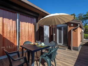 En udendørs terrasse med et bord, stole og en parasol, samt en lille bygning med dør og vindue.