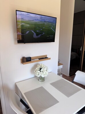 Ein flachbildfernseher an der Wand, der eine Landschaft zeigt, ein kleines Holzregal mit einer Uhr, eine Vase mit weißen Blumen und ein weißer Tisch mit Unterlage.