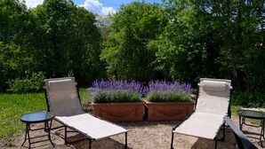 En utomhusplats med sittmöbler, bestående av två fåtöljer och ett litet bord, omgiven av grus och krukväxter med lila blommor.