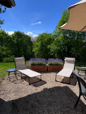 Ein Sitzbereich im Freien mit zwei Lounge-Stühlen und einem kleinen Tisch, umgeben von Kies und Blumentöpfen mit violetten Blüten.