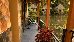 A stone-tiled balcony with a potted plant, a pair of green lounge chairs, and a view of a water body with thatched-roof structures. - Hang Mua Peaceful Retreat (Hoa Lu)