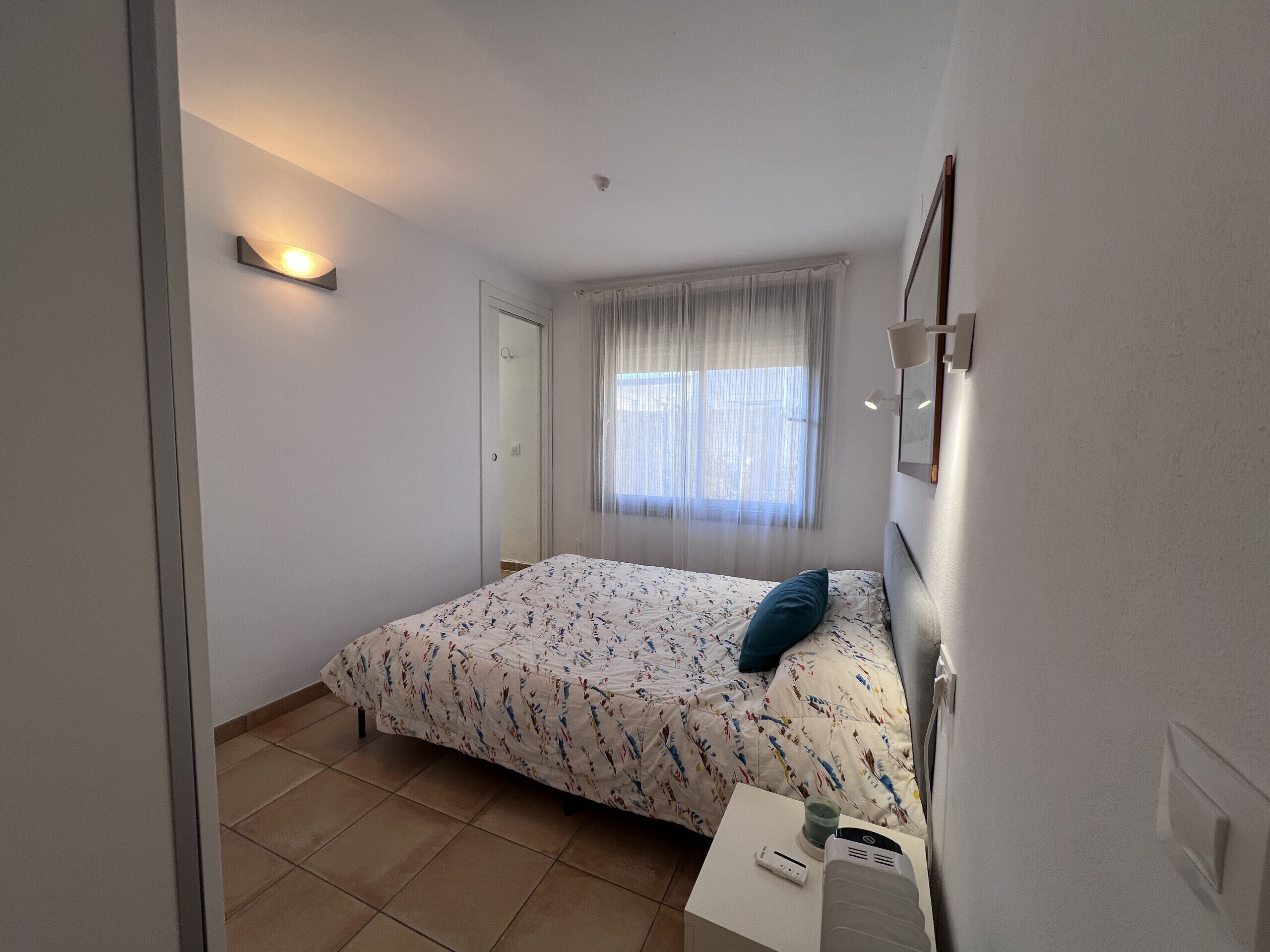 Ein Schlafzimmer mit Bett, Nachttisch, einem Fenster mit durchsichtigen Vorhängen und einer Wandleuchte.