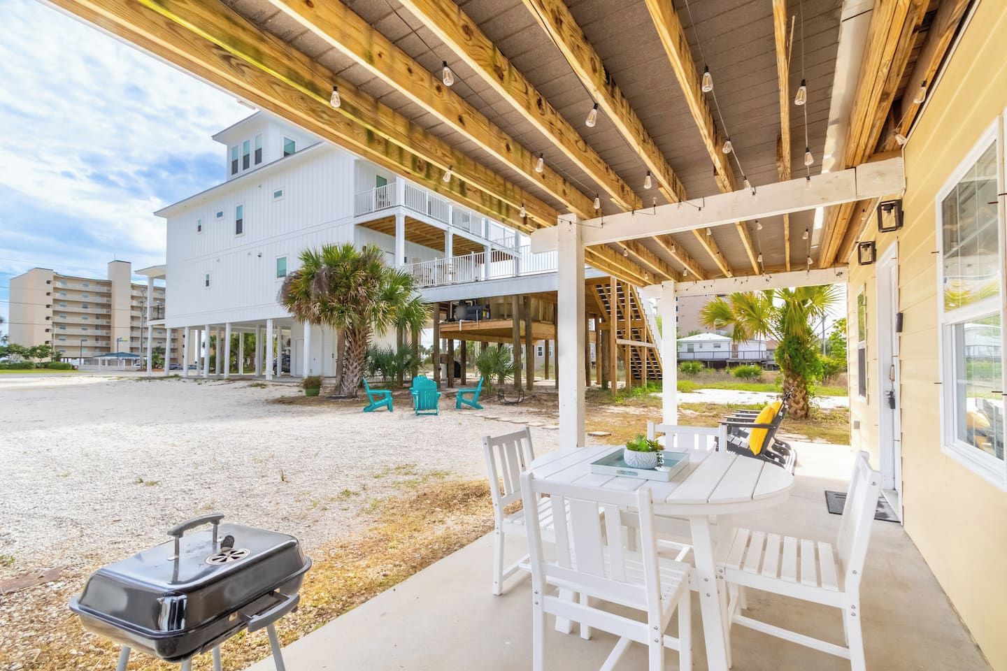 A covered outdoor seating area with a grill, white furniture, and a view of a multi-story building with balconies.