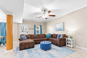 A living room with a sectional sofa, a ceiling fan, a blue ottoman, a small white side table, and a blue and white rug. - Multi-Family Home | Short Walk to Beach (Gulf Shores)