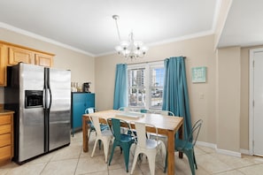 A kitchen with a stainless steel refrigerator, wooden cabinets, a dining table with chairs, a chandelier, and a window with curtains. - Multi-Family | Short Walk to Beach (Gulf Shores)