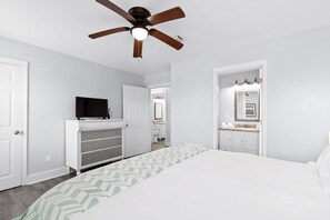 Ein Schlafzimmer mit Bett, Schminktisch, Fernseher, Deckenventilator und einem durch eine offene Tür einsehbaren Badezimmer.