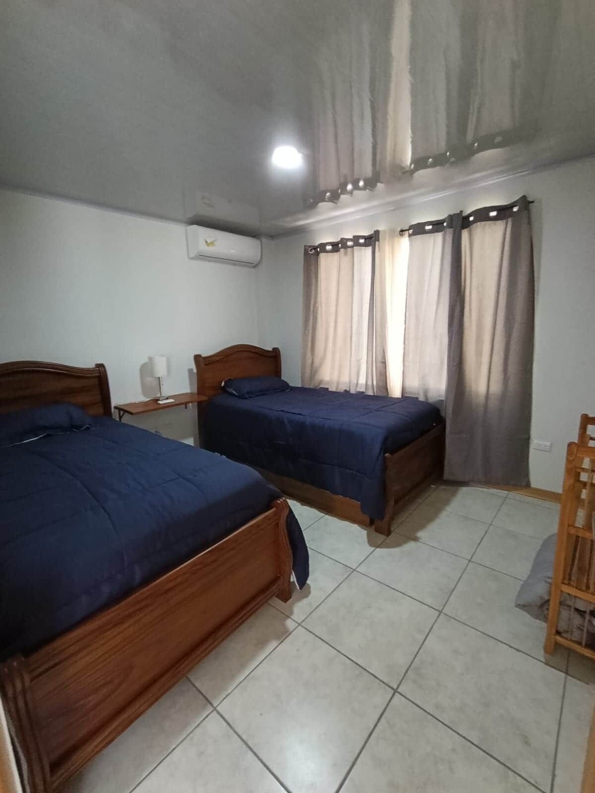 A bedroom with two wooden beds, a ceiling fan, a window with curtains, and a tiled floor.