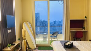 A hotel room with a bed, a desk, a chair, a TV, a balcony with a city view, and an air conditioning unit.