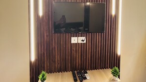 A flat-screen TV mounted on a wooden wall with built-in lighting, a shelf below holding remote controls, and small potted plants.