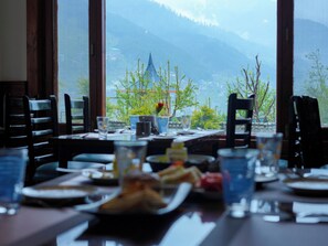 A dining table with chairs, plates, glasses, and a view of mountains through large windows. - The Radiant Resort Manali (Manali)