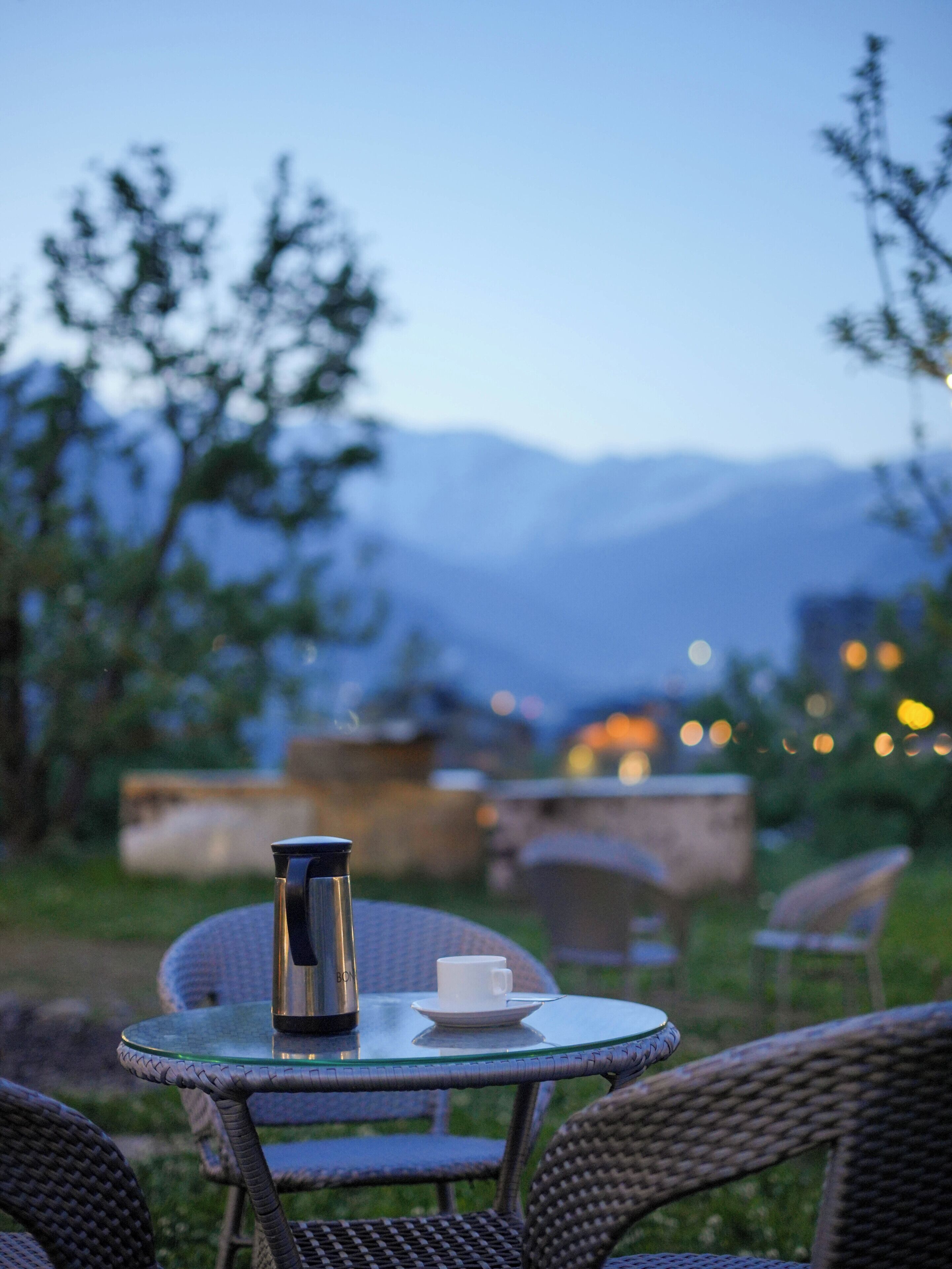 Una mesa de vidrio con una botella de agua de acero inoxidable y una taza blanca, rodeada de sillas de mimbre, ubicada al aire libre con vista a la montaña.