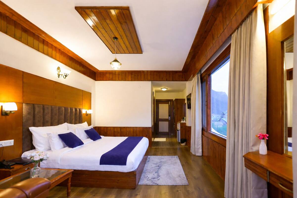 A hotel room with a wooden ceiling, a large bed with white linens, a bedside table, a nightstand, a window with curtains, and a view of the mountains.