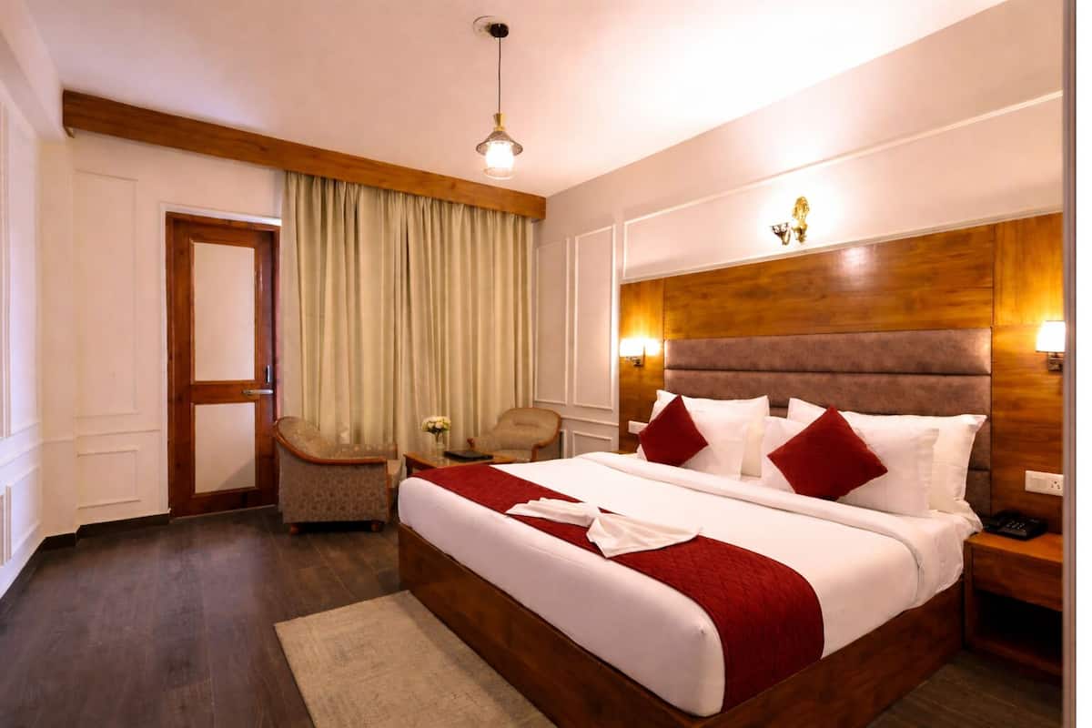 A hotel room with a large bed, a wooden headboard, two bedside tables, a nightstand, a desk, a chair, a lamp, and a window with curtains.