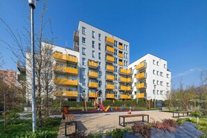 A multi-story residential building with balconies, a playground, and a paved area with benches.
