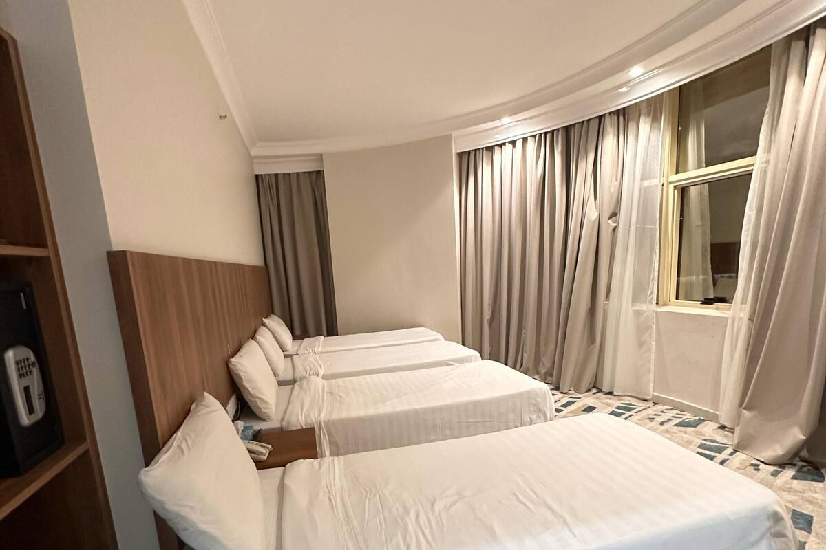 A hotel room with three beds, a wooden headboard, a bedside table with a phone, and a window with curtains.