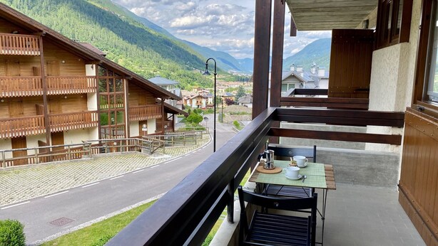 Un balcón con mesa y sillas, con vista a una calle y edificios con balcones de madera, al pie de montañas.