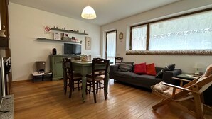 Una sala de estar con una mesa de comedor, sillas, un sofá, un televisor y una ventana con cortinas.