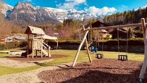 Un parque infantil con columpios y un tobogán, rodeado por un muro de piedra y una casita de juegos de madera.