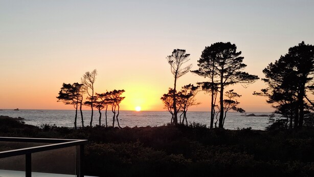 A sunset view from a balcony overlooking the sea with trees in the foreground. - Landrellec plage, large 4-bedroom house, sea view, piano, quiet location. (Pleumeur Bodou)