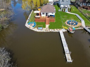 Ein Haus mit einer Anlegestelle, die sich über ein Gewässer erstreckt, umgeben von einem Rasen und einer Terrasse mit Sitzgelegenheiten.
